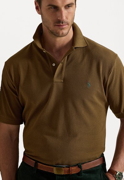 Camisa polo castanha com um acabamento texturizado, com botão na pala e um logo verde no lado esquerdo do peito. Combinada com um cinto castanho e um relógio.
