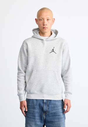 Szürke kapucnis pulóver, puha anyagból készült, elülső kenguruzsebbel és fekete Jumpman logóval a bal mellen. Farmernadrág van rajta.