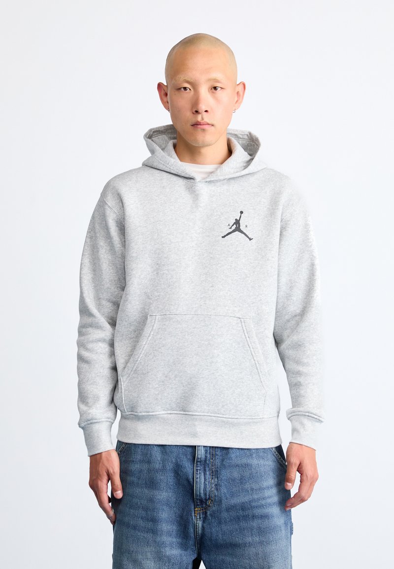 Felpa grigia con cappuccio realizzata in morbido tessuto, dotata di tasca frontale a marsupio e un logo Jumpman nero sul lato sinistro del petto. Indossati sotto, jeans in denim.
