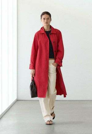 Kvinde går indendørs iført en lang rød frakke, sort top, cremefarvede bukser, sorte sandaler og holder en lille sort håndtaske i venstre hånd.