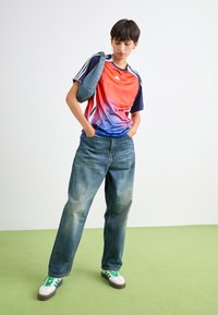 Sportiška trumpomis rankovėmis marškinėliai pereina iš raudonos į mėlyną, derinami su laisvo kirpimo išblukusiomis mėlynomis džinsomis ir žaliais raišteliais baltais sportbačiais.