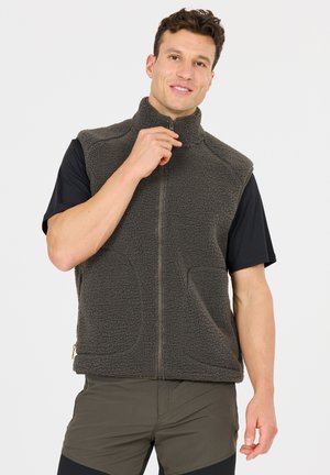 Mann trägt dunkelgraue Fleeceweste und schwarzes Kurzarmshirt, zieht den Kragen hoch, steht vor schlichtem hellem Hintergrund.