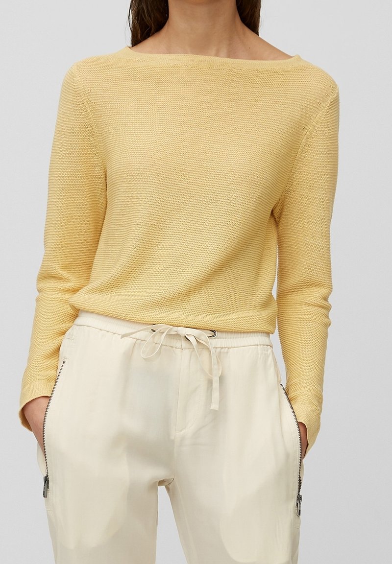 Femme portant un pull en maille jaune clair à manches longues et col bardot, associé à un pantalon crème à cordon avec poches zippées.