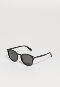 Ochelari de soare rotunzi negri cu lentile închise, având accente aurii și un detaliu cu dungi multicolore pe braț. Branding Polaroid observat.