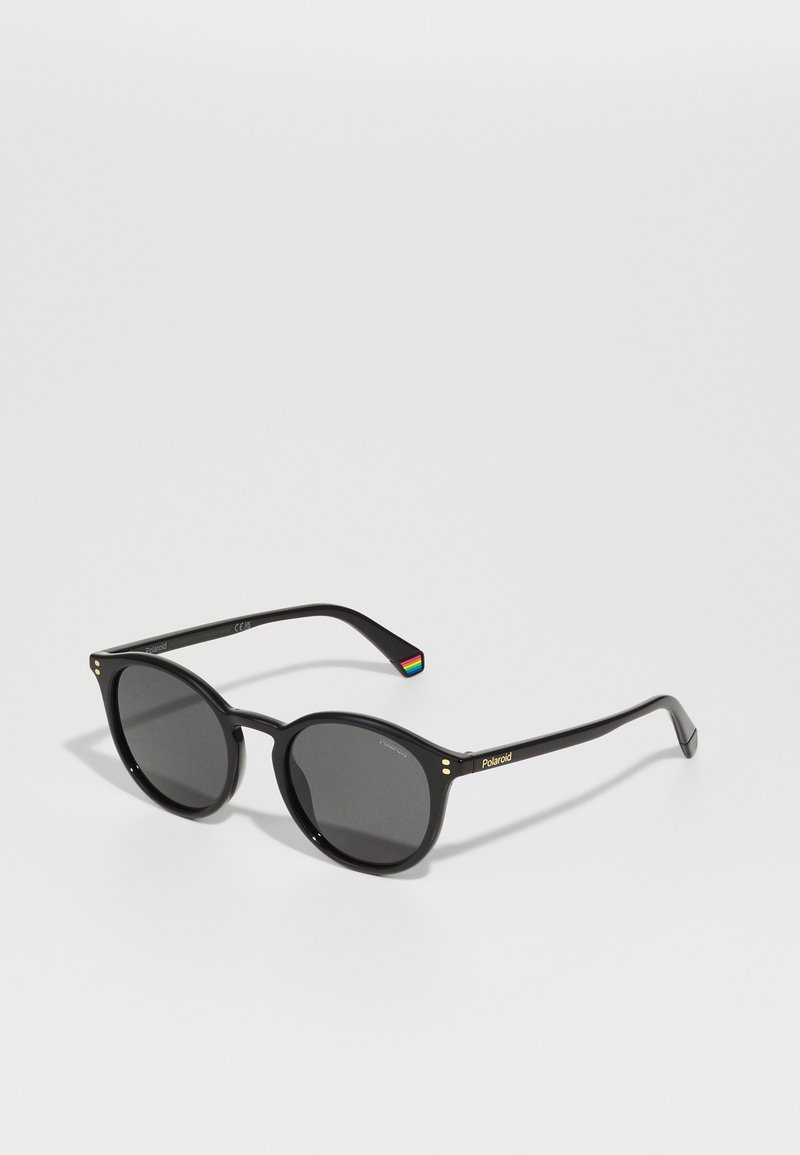 Ochelari de soare rotunzi negri cu lentile închise, având accente aurii și un detaliu cu dungi multicolore pe braț. Branding Polaroid observat.