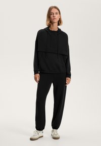 Felpa nera con collo alto, cordini e tasca frontale, abbinata a pantaloni jogger neri coordinati. Tessuto morbido con vestibilità rilassata. Sneakers bianche.