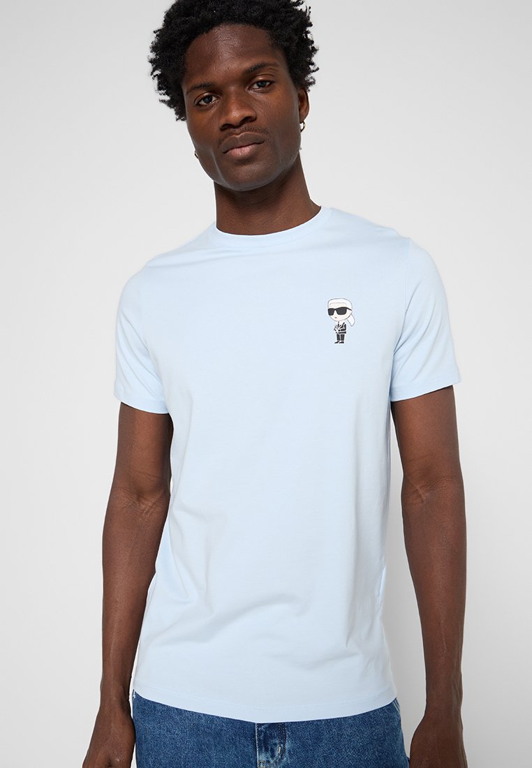 Hombre con camiseta azul clara con un pequeño gráfico de una figura de dibujos animados con gafas de sol en el pecho izquierdo, combinado con vaqueros azules.