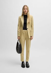 Jasnożółty garnitur skrojony na miarę z marynarką zapinaną na jeden guzik i prostymi spodniami, zestawiony z czarną bluzką i torebką. Czarne buty na platformie.