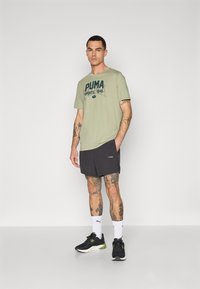 Svijetlo maslinasto zelena majica s tamnim printom, uparena s crnim šortsevima. Model nosi crne tenisice i bijele sportske čarape. Jednostavan dizajn.