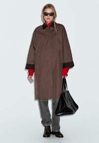 Cappotto oversize marrone con accenti neri, maglione rosso, jeans grigi e borsa nera. Presenta maniche larghe e tasche laterali.