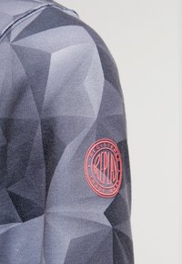 Sweatshirt gris avec un motif géométrique aux nuances variées et un patch logo circulaire rouge sur la manche gauche. Texture de tissu douce.