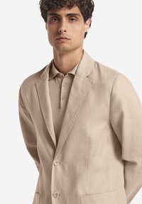 Beige linneblazer med slag, enkelknäppt design, två knappar och en subtil textur, kombinerad med en matchande pikétröja.