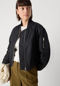 Giacca bomber nera cropped con motivo trapuntato, chiusura con zip argentata, colletto a coste e tasca con zip sulla manica; tessuto leggero e morbida al tatto.