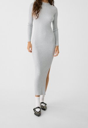 Kvinde iført en langærmet, ribbet lys grå midi-kjole med sideslidser, hvide sokker og sorte Mary Jane-sko med nitter, stående på gråt gulv.