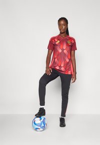 Raudona sportinė marškinėlė su abstrakčiu raštu, juodos kelnės ir juodi batai, stovi ant spalvingo futbolo kamuolio.