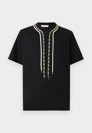 Schwarzes T-Shirt mit kurzen Ärmeln und zwei langen dekorativen Kordeln, eine beige und eine grün gemustert, die vom Kragen hängen.