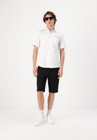 Camisa blanca de manga corta con botones y un bolsillo, acentuada con detalles en azul, combinada con pantalones cortos negros y zapatillas blancas. Textura suave, ajuste casual.