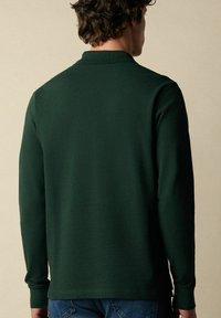 Poloko s dlouhým rukávem ve tmavě zelené barvě z bavlny. Obsahuje límec a žebrované manžety. Zadní pohled ukazuje čistý, minimalistický design.