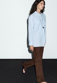 Donna che cammina indossando una camicia azzurra chiaro con bottoni, pantaloni larghi marroni, sandali neri, una lunga collana e anelli, su uno sfondo bianco semplice.