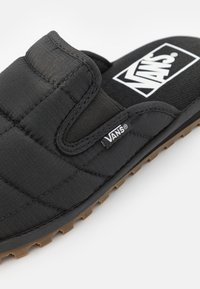Svart quilted slip-on sko med polstret overdel, teksturert fotbed og gummisåle med små mønstre. Vans-logo på siden.