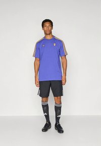 Camisola atlética roxa com riscas douradas, calções pretos e meias pretas até ao joelho com design de caveira, usadas com chuteiras de futebol pretas.