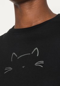 Camiseta negra de algodón con un sencillo diseño de cara de gato gris claro, con bigotes, presentado en una textura suave.