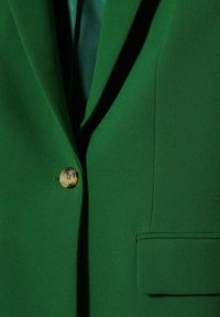 Gros plan sur un blazer vert avec un seul bouton marron boutonné, doté d'un revers cranté et d'une poche à rabat sur le côté droit.