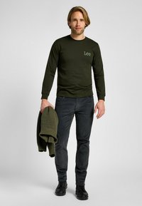 Camicia a maniche lunghe di colore verde oliva con un piccolo logo ricamato "Lee", abbinata a pantaloni grigio scuro e a un maglione in maglia coordinato appoggiato sul braccio.