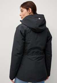 Zwarte waterdichte jas met een aangesnoerde taille, verstelbare capuchon en een logodetail op de achterkant. Gladde, matte textuur.