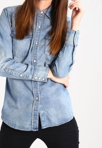 Ljusblå denimskjorta med knäppning framtill, två bröstfickor, långa ärmar och knäppta manschetter. Har en rundad nederkant och lätt fransning.