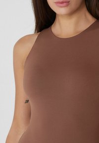 Mujer con cabello largo castaño, vistiendo una blusa marrón sin mangas, con un pequeño tatuaje de corazón visible en su brazo izquierdo superior.