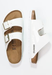 Sandalias Birkenstock blancas con dos hebillas ajustables, plantilla de corcho y suela de goma texturizada, con un acabado brillante y detalle del logo.