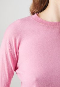 Jasnoróżowy sweter dzianinowy z gładką fakturą, okrągłym dekoltem i długimi rękawami. Materiał wydaje się miękki i dopasowany w okolicy ramion.