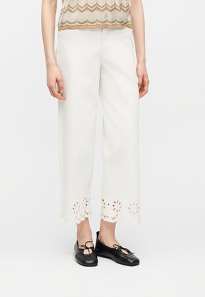 Femme portant un pantalon blanc court avec un ourlet découpé floral et des ballerines noires, debout devant un fond uni.