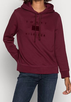 Personne portant un sweat à capuche bordeaux Tommy Hilfiger avec cordons et un jean bleu foncé, debout avec une main sur la hanche devant un fond uni.