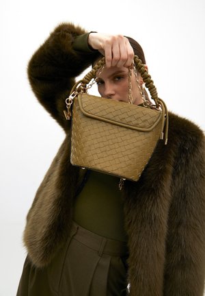 Borsa a tracolla in pelle intrecciata beige con dettagli in catena dorata e manico annodato, con chiusura a pattina e una texture liscia, tenuta contro una pelliccia grigia.