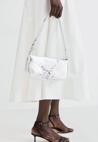 Borsa a mano metallica argentata con design texturizzato e dettaglio decorativo con lacci. Presenta una sottile tracolla e una forma rettangolare strutturata.
