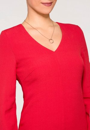 Femme portant un haut rouge à col en V et un collier doré avec un pendentif circulaire, visible des épaules au menton.