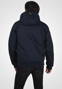 Marineblauwe gewatteerde jas met een capuchon, elastische manchetten en zoom. Bevat een klein leren label aan de achterkant. Zichtbare stikseldetails.