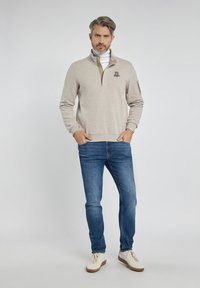 Beżowy sweter z ćwiartkowym zamkiem, haftowanym logo po lewej stronie klatki piersiowej, w połączeniu z niebieskimi dżinsami i białymi trampkami. Gładka tekstura.