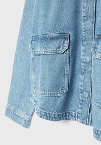 Manche de veste en denim bleu clair avec grande poche plaquée avant et coutures visibles sur fond uni.