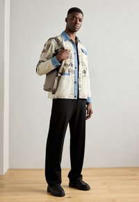 Beige Jacke mit blauem Denim-Kragen, grafischen Druckmotiven, über einem blauen Hemd getragen. Schwarze Hose und klobige schwarze Schuhe, stehend auf einem Holzboden.