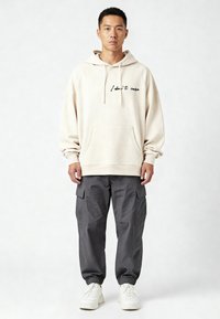 Felpa beige con scritta "Non me ne importa", tasca a marsupio frontale, abbinata a pantaloni cargo grigi con tasche laterali e sneakers bianche.