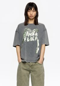 Camiseta gris de gran tamaño hecha de algodón, con una textura desgastada y una impresión gráfica metálica verde que dice "Bimba y Lola."