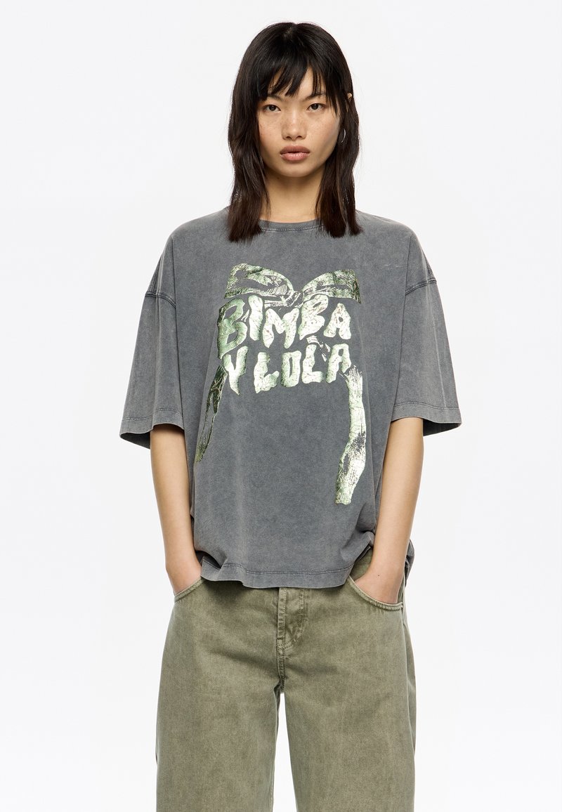 Camiseta gris de gran tamaño hecha de algodón, con una textura desgastada y una impresión gráfica metálica verde que dice "Bimba y Lola."