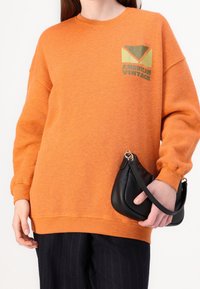 Oranssi collegepaita, jossa on pyöreä pääntie ja laskostetut olkapäät, jossa on graafinen painatus, jossa lukee "AMERICAN VINTAGE". Musta käsilaukku, jossa on kaareva muoto.