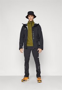 Chaqueta impermeable negra con detalles grises, forro polar verde oliva debajo, pantalones negros y zapatos de senderismo naranjas, complementado con un sombrero de cubo negro.