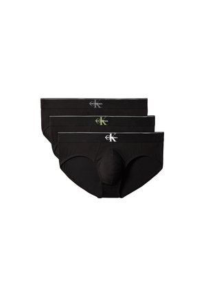 Homme musclé debout pieds nus devant un fond gris, portant un slip noir et tenant la taille avec les deux mains.