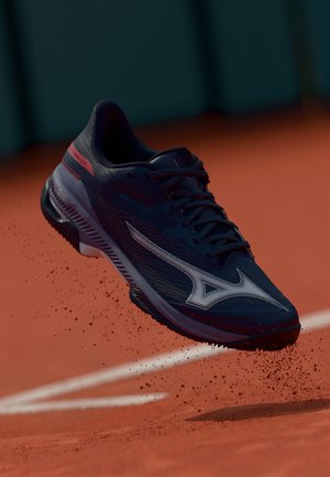 Blue athletic shoe with white accents, textured surface, and pronounced lacing system, suspended above a red clay court with dust.