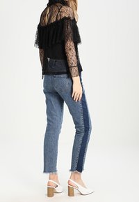 Kvinde iført en gennemsigtig sort blonder-top med volanger, blå to-tonede jeans med rå kanter og hvide blokhælede slingback-sandaler, stående sidelæns.
